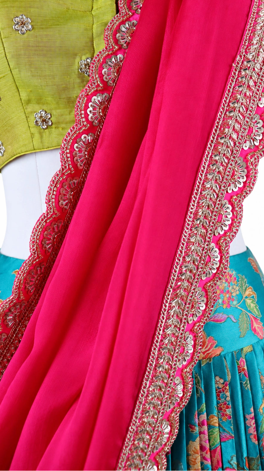 Turquoise florals, fresh green and bright pink bring a vivid contrast to this three-piece lehenga set by Vinisha Rao Designer Studio. The gathered Gaji silk lehenga features multicoloured floral motifs and a broad pink patterned border. The green raw silk blouse has a front button fastening, elbow-length sleeves and metallic floral embroidery around the neckline and cuffs. An open back with tassel ties completes the blouse, while the pink chiffon dupatta adds a soft layer with an embellished scalloped borde